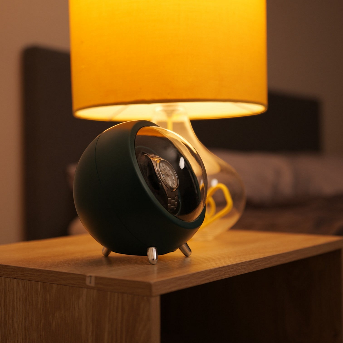 Winderly Cyclops watch winder vert foncé avec couvercle transparent sur une table de chevet près d'une lampe